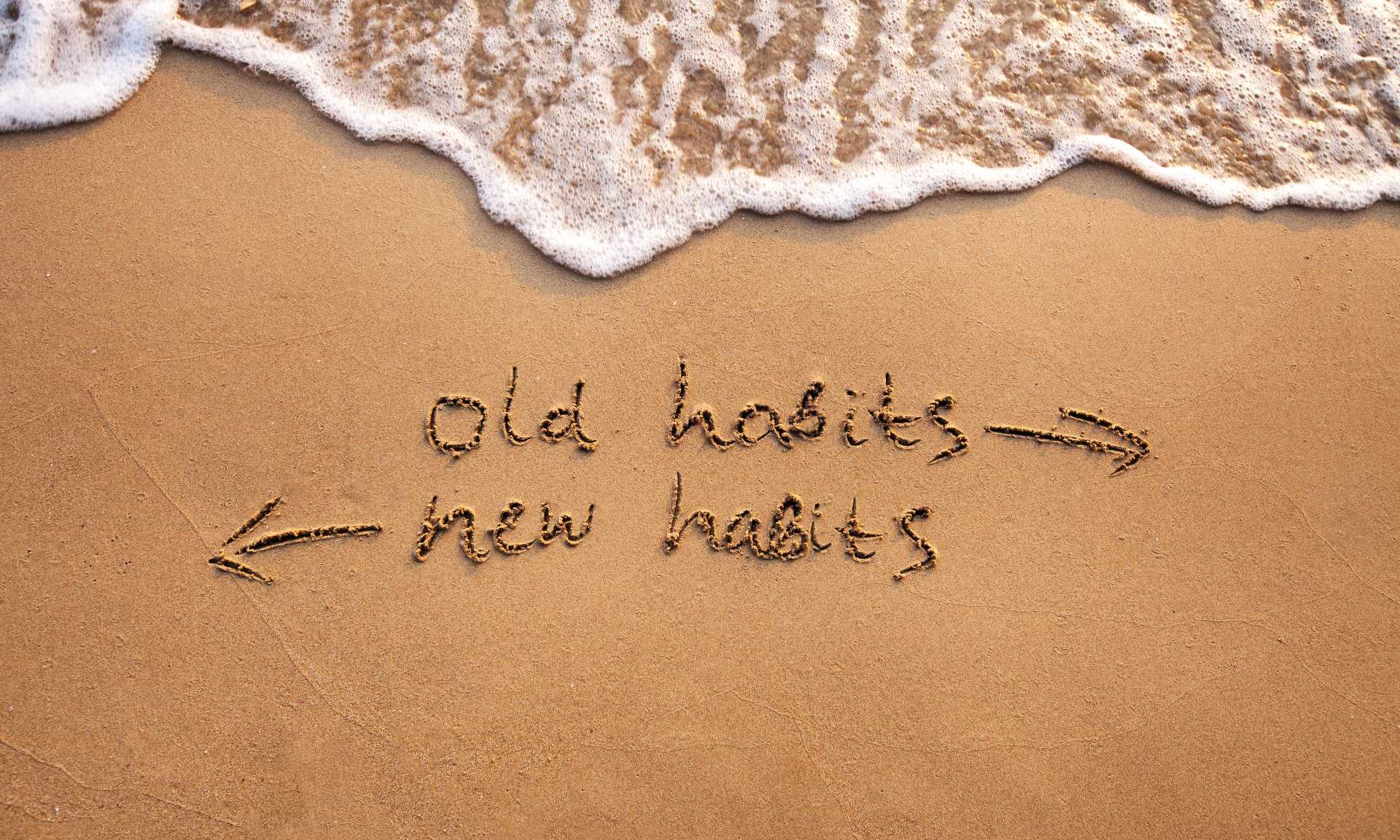 The words old habits and new habits written in beach sand with arrows pointing in either direction and the water approaching