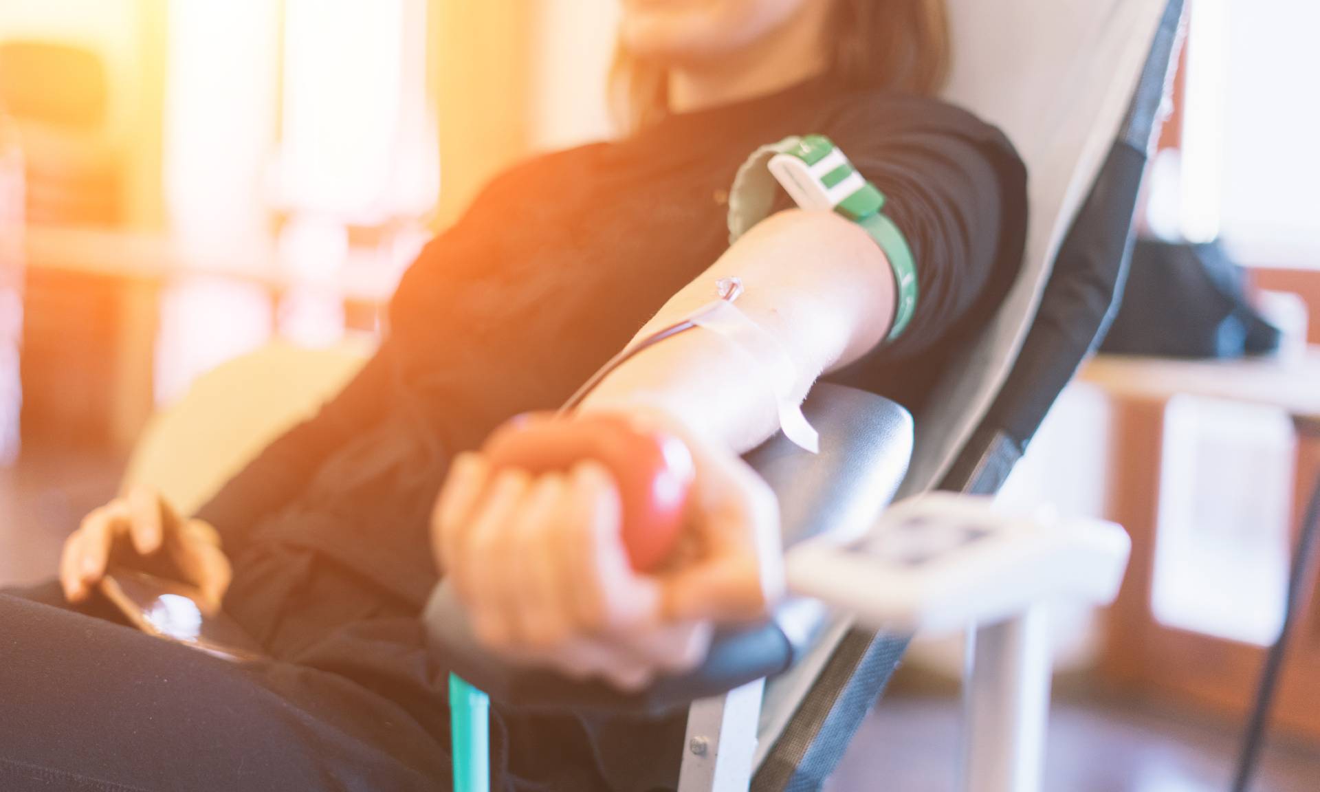 Young woman with arm outstretched and hand squeezing small red rubber ball who is donating blood. 