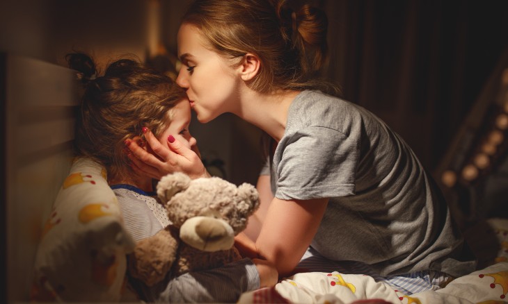 Mother kisses young daughter on the head while the girl lies in bed holding her teddy bear.