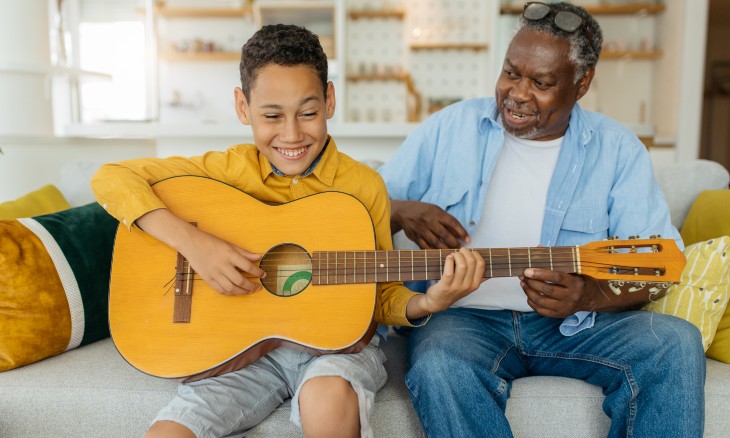 Young boy strumming a guitar while sitting next to his grandfather.