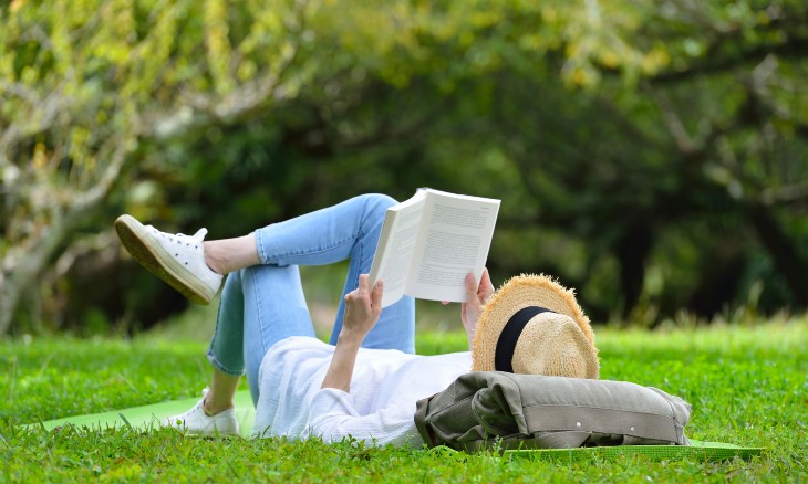 Woman with sunhat laying on a blanket in a park and reading a book